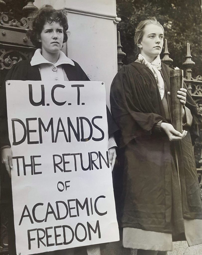 A woman holds a placard that says "UCT demands the return of academic freedom" beside a woman holding a lit torch. Both are in academic dress.